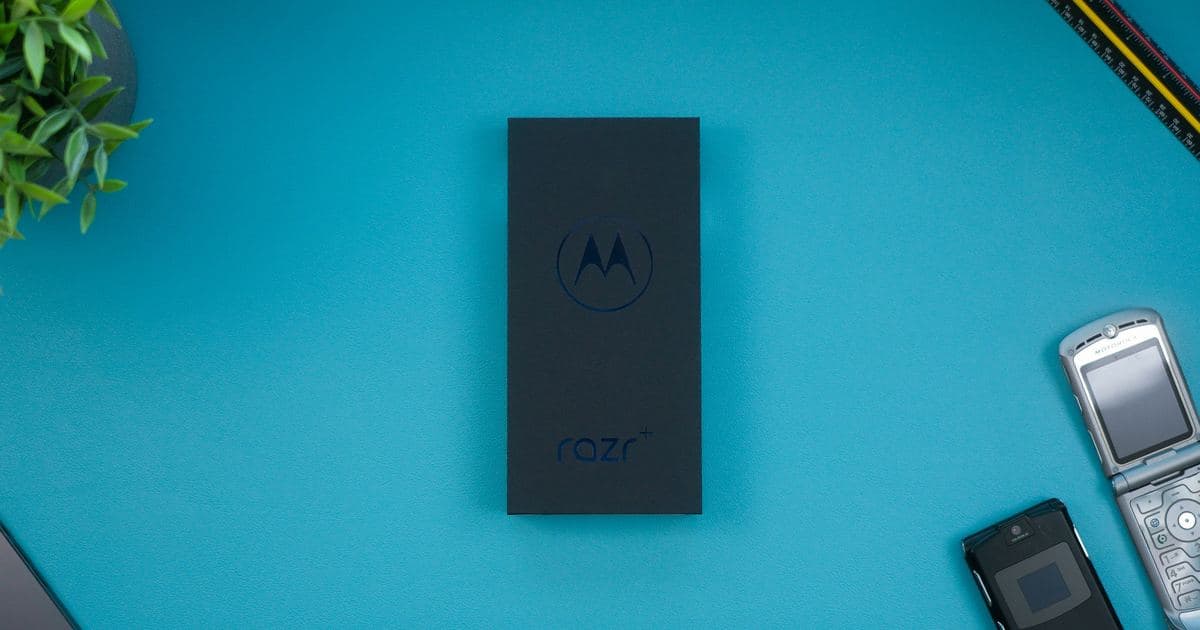 Caixa do smartphone dobrável Motorola Razr+ sobre uma mesa azul, com o logo da Motorola em destaque e dois aparelhos Motorola RAZR mais antigos ao lado.