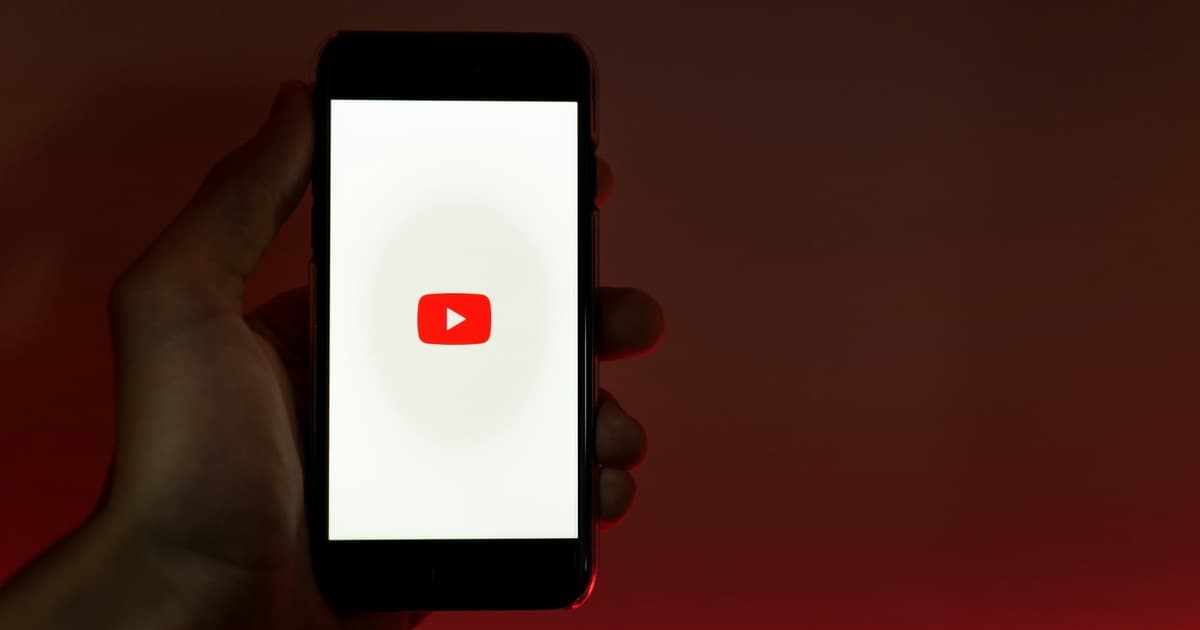 Mão segurando um celular com o logotipo do YouTube na tela, contra um fundo escuro avermelhado.