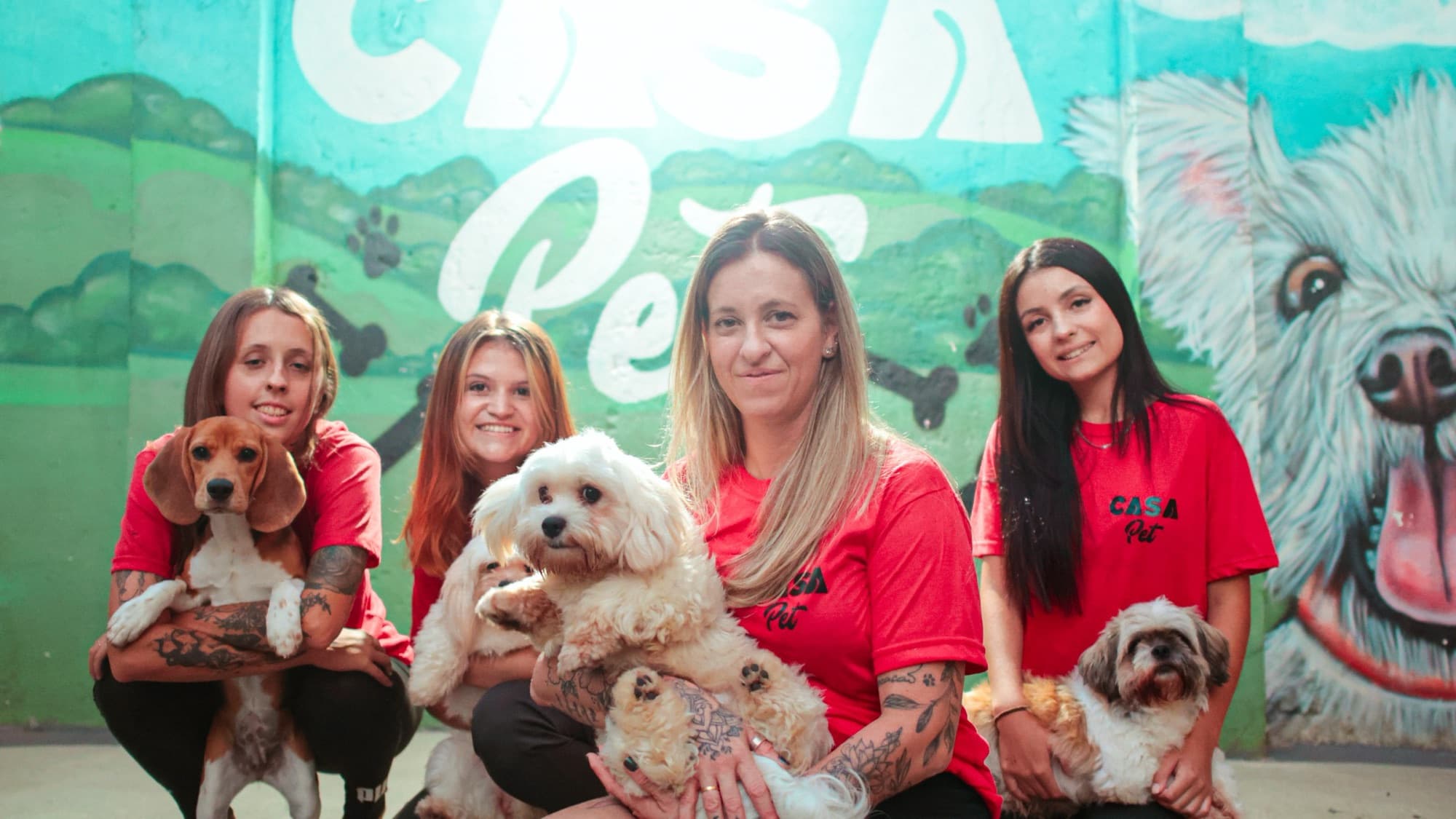 As quatro tias da Casa Pet agachadas com os cães no colo, diante do mural pintado com o letreiro Casa Pet