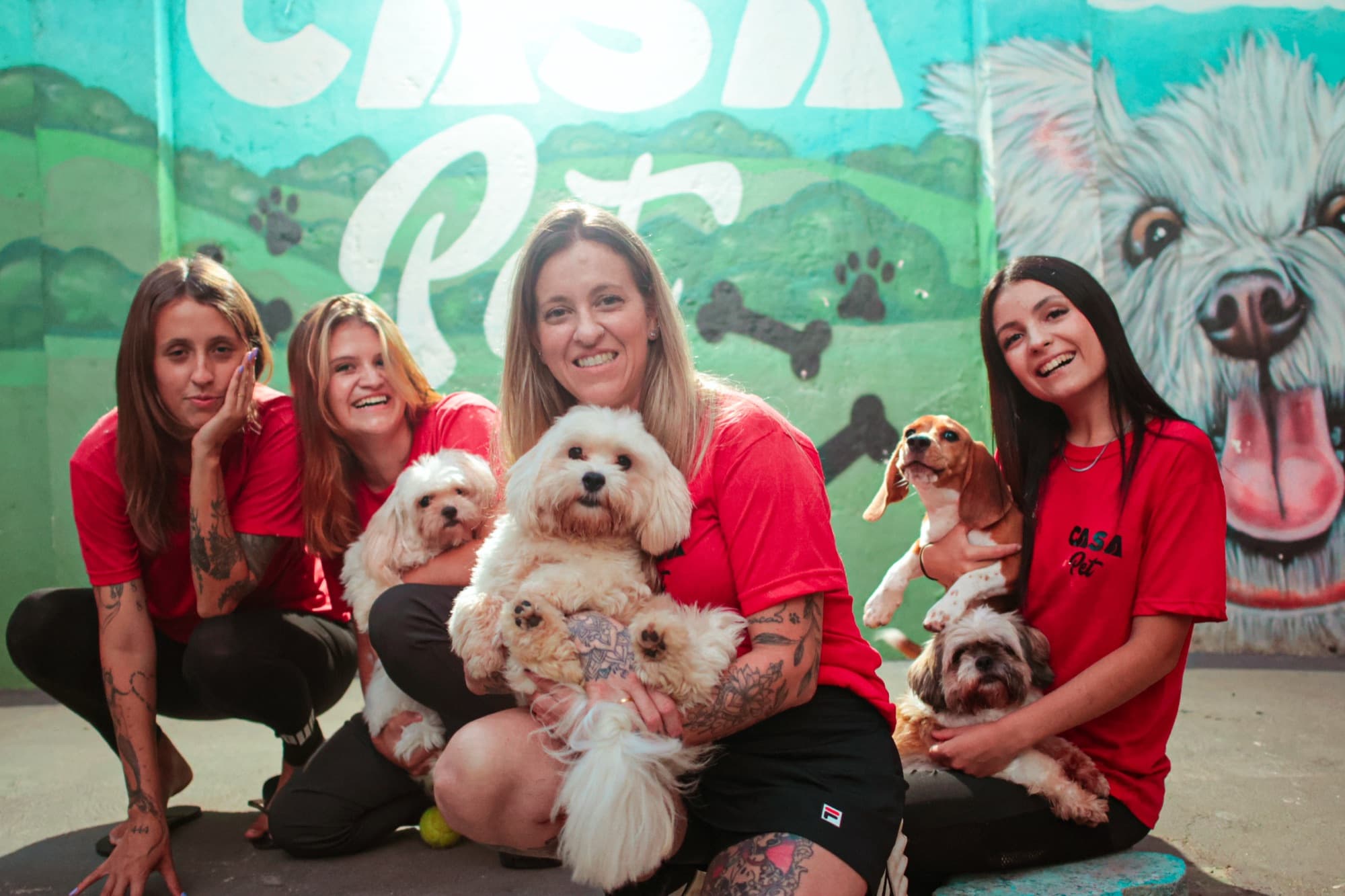 As tias da Casa Pet agachadas com cães no colo, em frente ao mural pintado com paisagem e cachorros