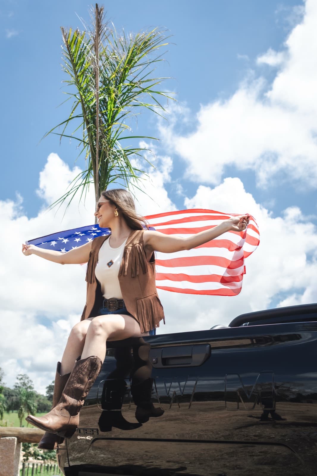 Demy Manosso sentada na caçamba da picape segurando a bandeira sob um céu azul
