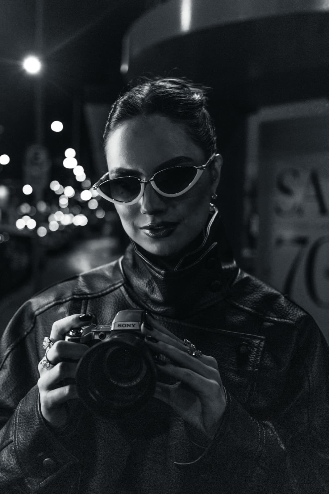 Retrato em preto e branco de Demy Manosso com óculos gatinho e câmera fotográfica na rua
