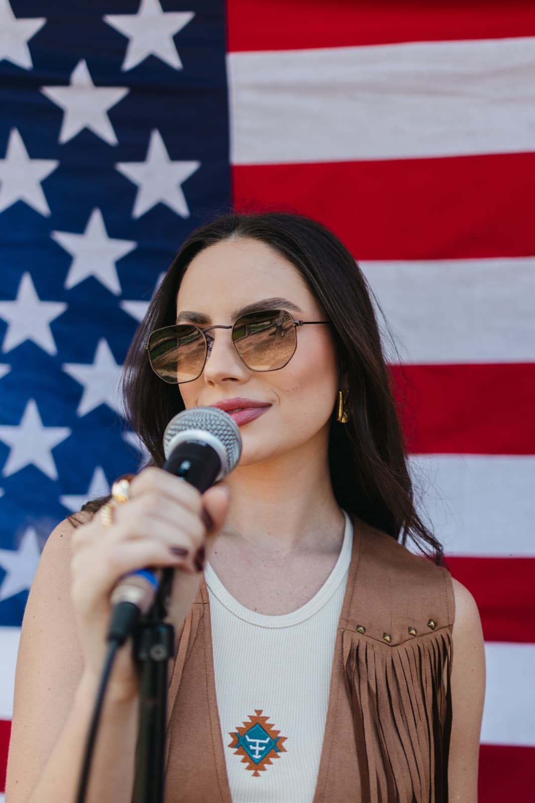 Demy Manosso canta ao microfone com óculos de lentes âmbar e colete de franja