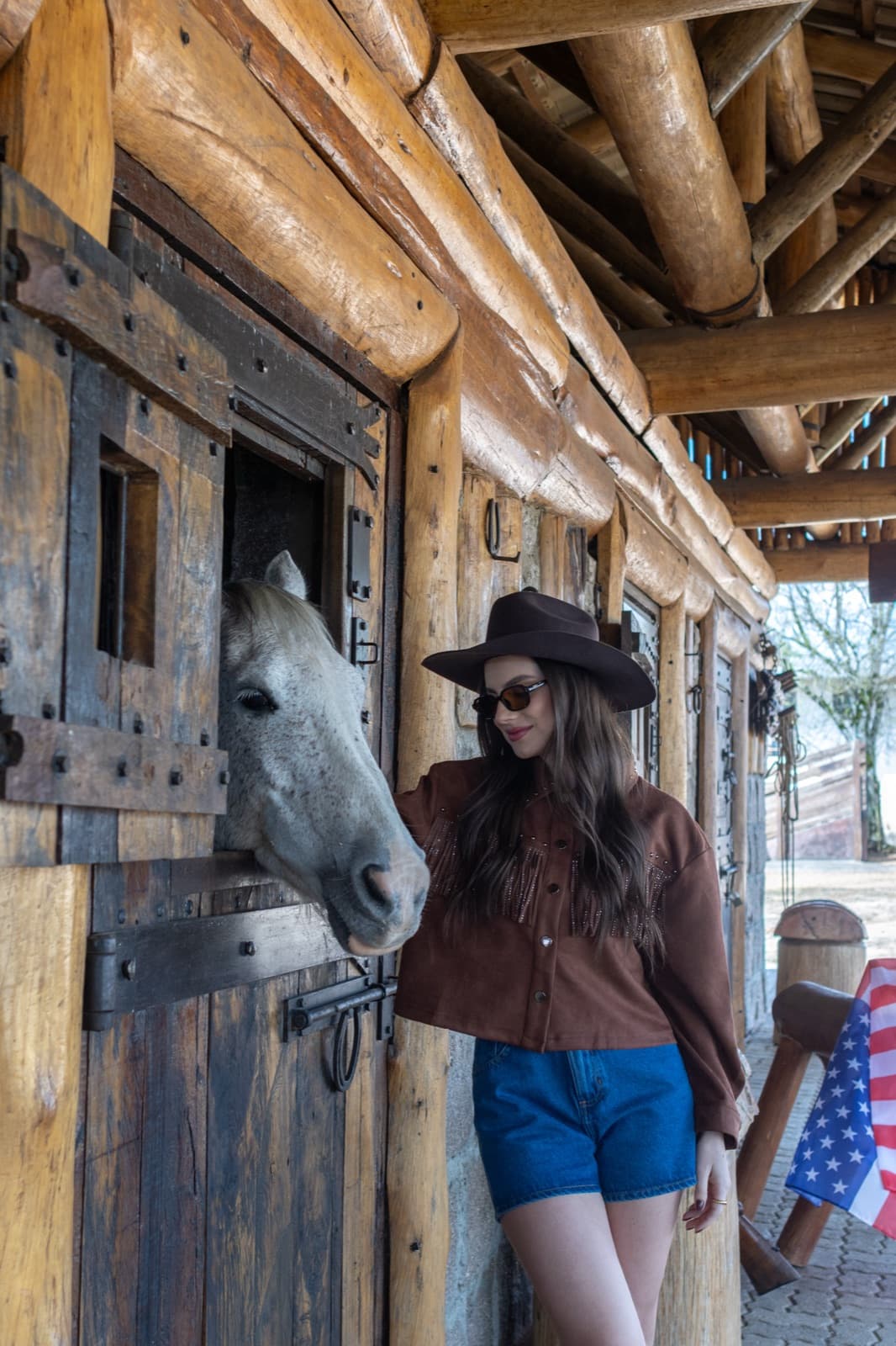Demy Manosso encostada na porta da baia, com o cavalo branco ao lado
