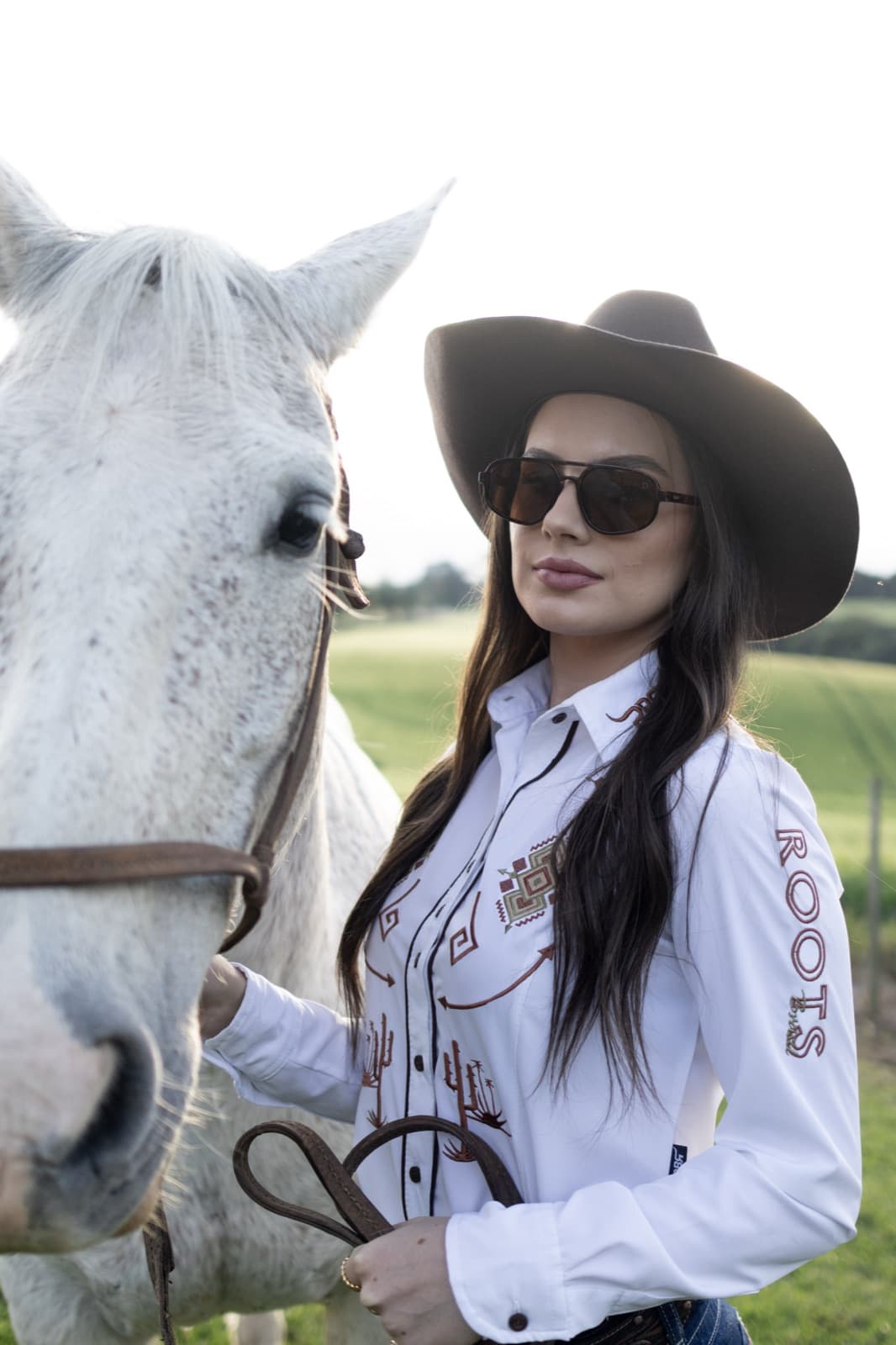 Demy Manosso de camisa branca bordada ao lado de um cavalo branco no pasto