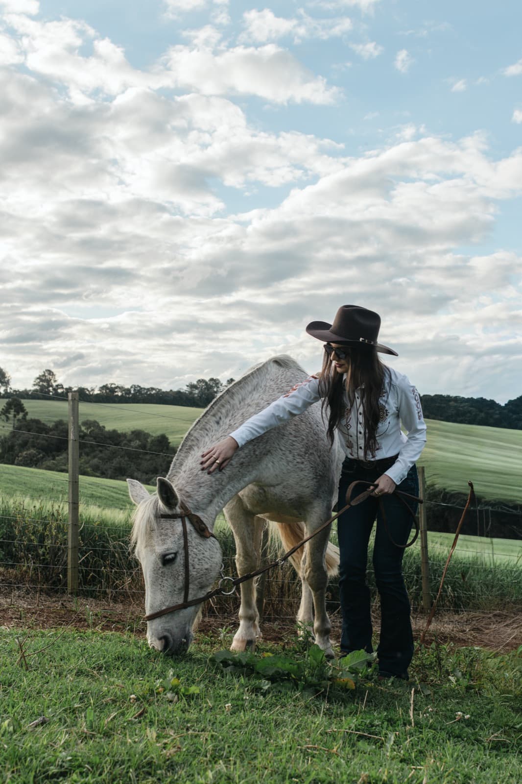 Demy Manosso acaricia um cavalo branco que pasta sob um céu de nuvens